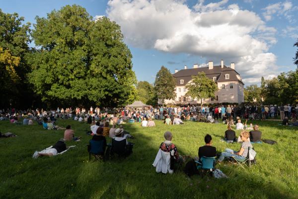 Viele Besucherinnen und Besucher sitzen und stehen auf einer Wiese vor einem hellen Schlossgebäude unter blauem Himmel.