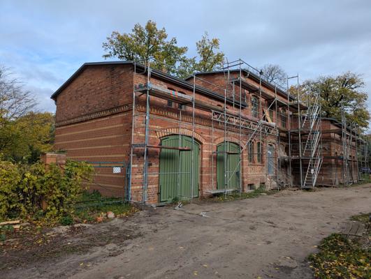 Ein langes Backsteingebäude steht unter Sanierung, umgeben von Gerüsten und mit grünen Toren an der Fassade.