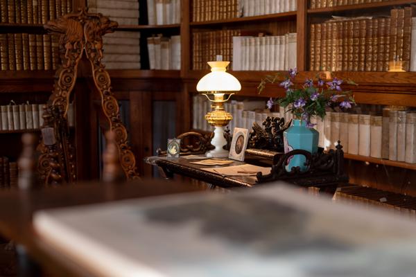 In einer historischen Bibliothek leuchtet eine Tischlampe über einem Schreibtisch mit Blumen, Büchern und kleinen Rahmen.
