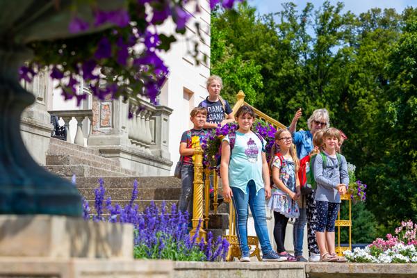 Mehrere Kinder und eine ältere Frau stehen auf den Stufen vor einem hellen Gebäude, umgeben von violetten Blumen.