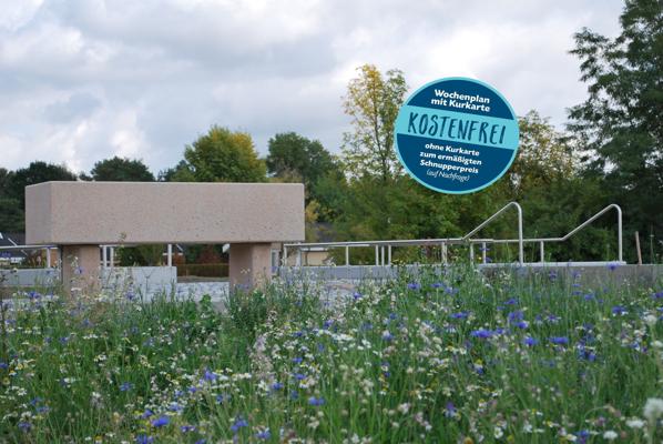 Ein rundes Schild nennt den Wochenplan mit Kurkarte und gratis Angebot, davor liegen ein Beckenrand und eine blühende Wiese.