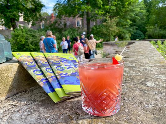 Ein rosafarbener Cocktail mit Fruchtspieß steht vor Pückler-Broschüren auf einer Steinmauer, dahinter gehen Menschen.