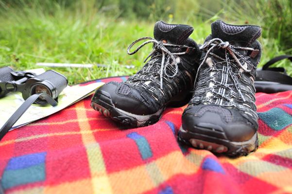 Schwarze Wanderschuhe liegen auf einer bunten Decke im Gras, daneben sind ein Fernglas und eine Karte zu sehen.