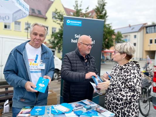 Drei ältere Personen stehen an einem Infostand des Weißen Rings und reichen sich Broschüren auf dem Wochenmarkt.