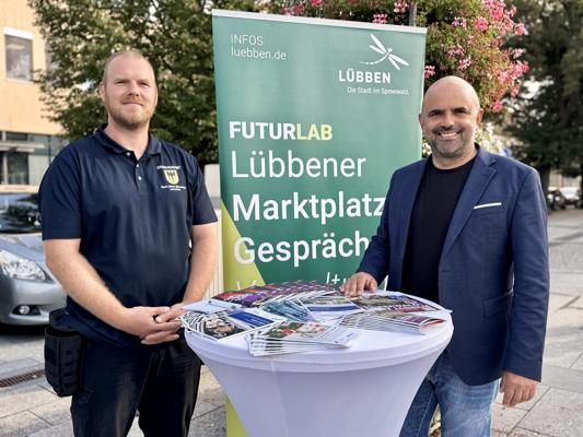 Zwei Männer stehen vor einem grünen Banner mit der Aufschrift LÜBBEN und FUTURLAB; auf dem Tisch liegen Flyer.