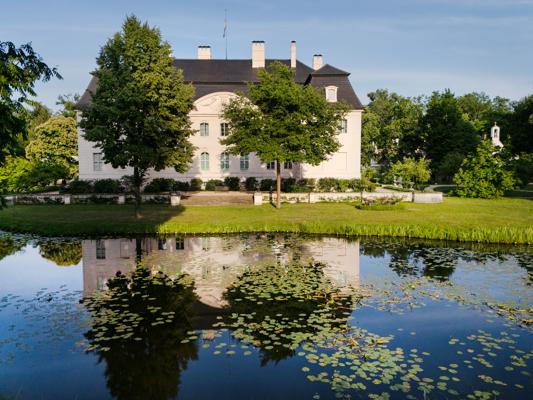 Ein weißes Herrenhaus steht hinter Bäumen am Ufer eines Teichs, dessen Wasser das Gebäude und Seerosen deutlich spiegelt.