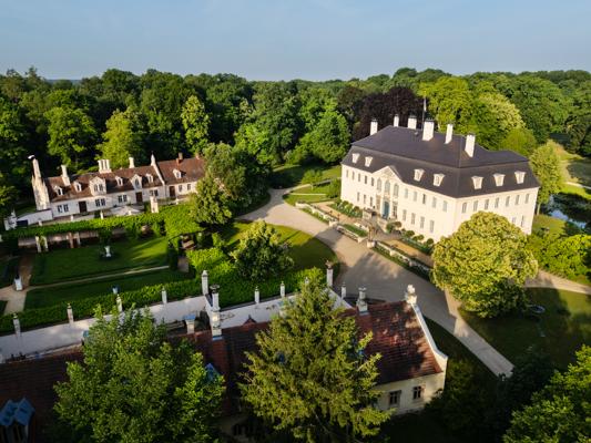 Aus der Luft sind ein helles Schlossgebäude, ein Seitenflügel und gepflegte Gartenanlagen mit Wegen und Baumbestand zu sehen.