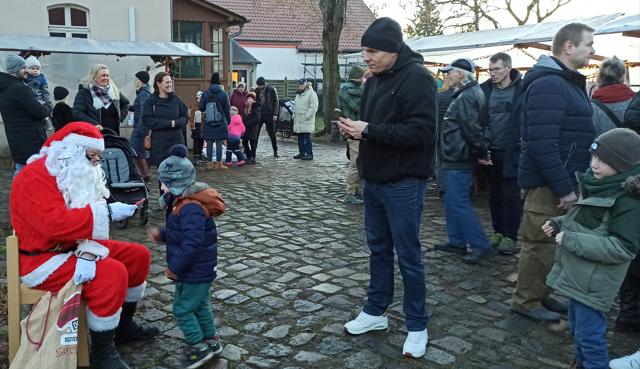 Auf dem gepflasterten Hof sitzt der Weihnachtsmann im roten Kostüm und spricht mit einem Kind, während Besucher vorbeigehen.