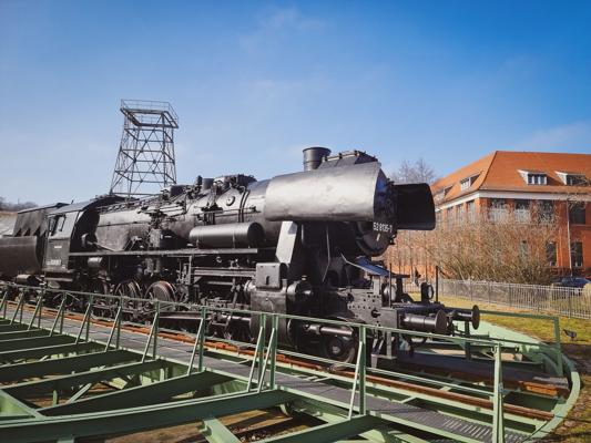 Eine schwarze Dampflokomotive steht auf Gleisen vor einem Fördergerüst und einem Backsteingebäude unter blauem Himmel.
