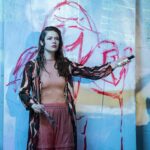 Eine Frau mit nassem Haar steht vor einer blauen Wand mit roter Kritzelei und blickt konzentriert zur Seite.