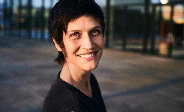 Eine kurzhaarige Frau lächelt in der Abendsonne vor einer modernen Glasfassade und blickt direkt in die Kamera.