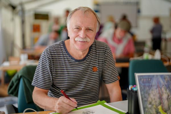 Ein lächelnder älterer Mann mit grauem Schnurrbart hält einen roten Bleistift am Zeichentisch in einer hellen Werkstatt.