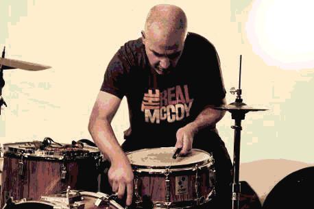 Ein glatzköpfiger Schlagzeuger spielt konzentriert auf der Snare, während auf seinem T-Shirt „The Real McCoy“ steht.