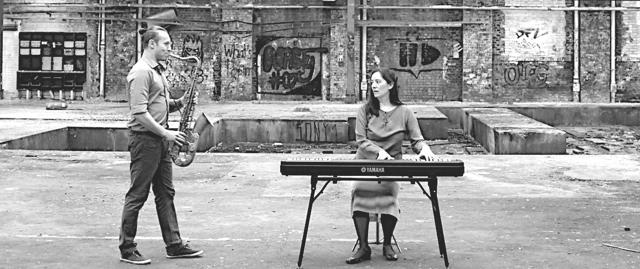 Ein Saxophonist steht vor einer sitzenden Keyboarderin in einem industriellen Hof mit Graffiti an der Backsteinwand.