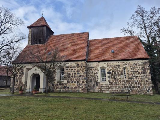 Feldsteinkirche mit hölzernem Turmaufsatz, rotem Ziegeldach und rundbogigem Eingang in einer grünen Wiese.