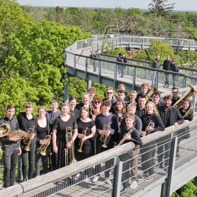 Eine Gruppe junger Musiker steht mit Trompeten und Posaunen auf einer geschwungenen Plattform über dichtem Grün.