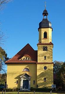 Eine gelbe Dorfkirche mit rotem Dach, hohem Turm und Eingangsvorbau steht unter klarem blauem Himmel.