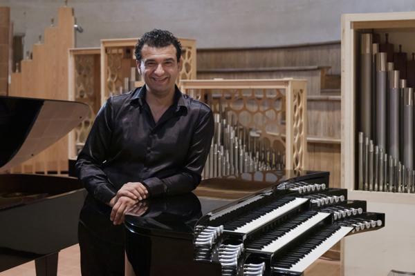 Ein lächelnder Organist sitzt an einer Orgel mit sichtbaren Pfeifen und Tasten in einem hellen Konzertsaal.