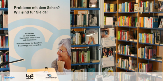 In einer Bibliothek steht ein Poster mit der Aufschrift „Probleme mit dem Sehen? Wir sind für Sie da!“ vor Bücherregalen.