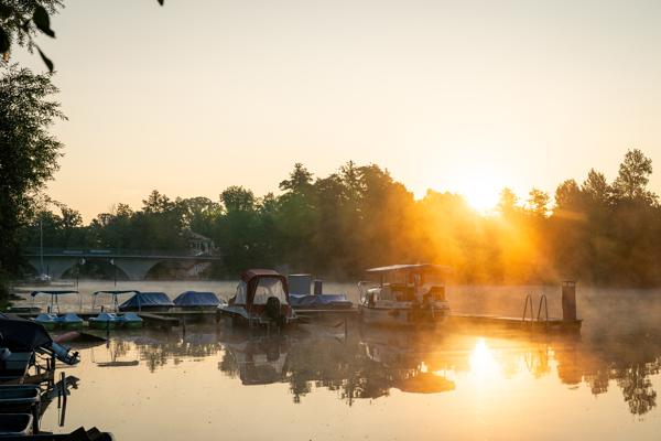 Boote liegen ruhig auf dem spiegelnden Wasser am Spreeufer, während die Sonne durch den Morgennebel aufsteigt.
