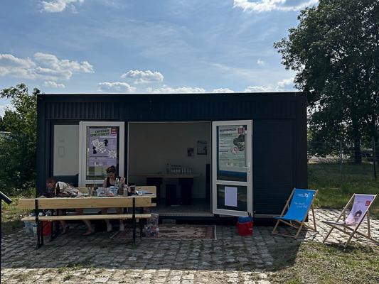 Vor einem dunkel verkleideten Container steht ein Basteltisch; daneben sind Klappstühle und Poster zu sehen.