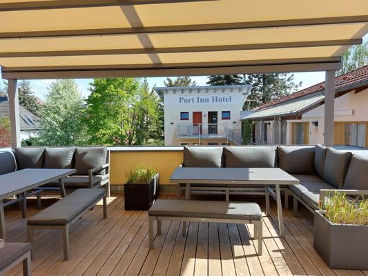 Auf der Holzterrasse des Port Inn Hotel stehen Sitzgruppen mit Sofas, Tischen, Bänken und Pflanzkübeln unter der Überdachung.