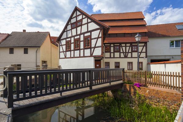 Ein Fachwerkhaus mit rotem Ziegeldach steht an einer kleinen Brücke über einem schmalen Wasserlauf in einer Altstadt.