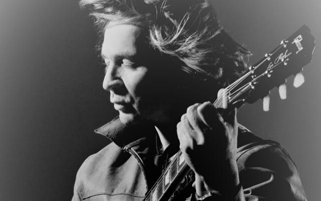 Ein Musiker hält eine Gitarre nah am Gesicht, das Schwarzweiß-Porträt zeigt konzentrierten Blick und dramatisches Licht.