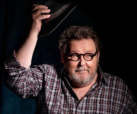 Ein bärtiger Mann mit runder Brille hebt einen dunklen Hut über den Kopf und blickt direkt in die Kamera.