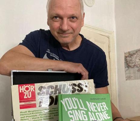 Ein älterer Mann hält zwei Bücher vor sich; auf den Covern stehen „You’ll Never Sing Alone“, „SCHUSS“ und „HÖRZU“.