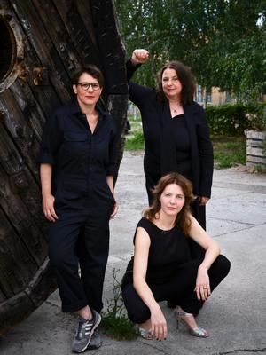 Drei Frauen stehen und sitzen vor einer großen Holzskulptur im Freien; eine hebt den Arm, zwei tragen dunkle Kleidung.
