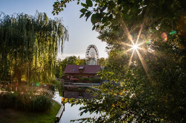 Zwischen Weidenzweigen und Ufergrün ragt ein Riesenrad hinter einem Haus auf, die Sonne glitzert durchs Laub über dem Wasser.