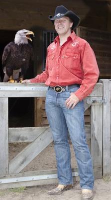 Ein Mann im schwarzen Cowboyhut und rotem Hemd lehnt am Holztor, neben ihm sitzt ein Weißkopfseeadler.