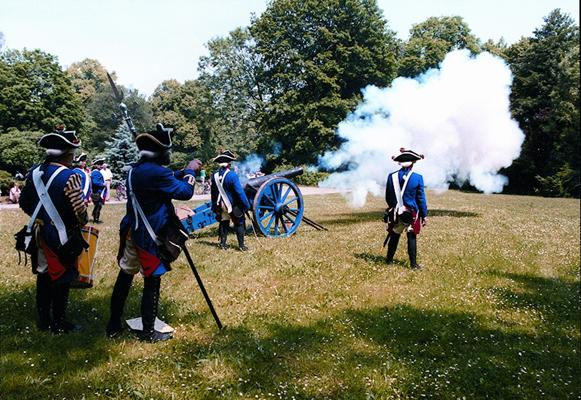 Mehrere Kanoniere in blauen Uniformen feuern eine Feldkanone auf einer Wiese ab, während dicker Rauch aufsteigt.