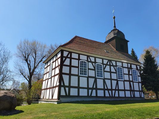 Eine weiß verputzte Fachwerkkirche mit rotem Ziegeldach steht auf einer Wiese, daneben ein dunkler Turm mit Wetterfahne.