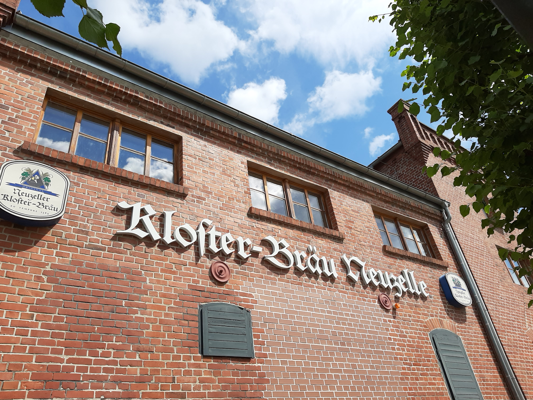 Die Backsteinfassade der Kloster-Brauerei Neuzelle zeigt große Fenster, das Schriftlogo und einen Blick in den Himmel.