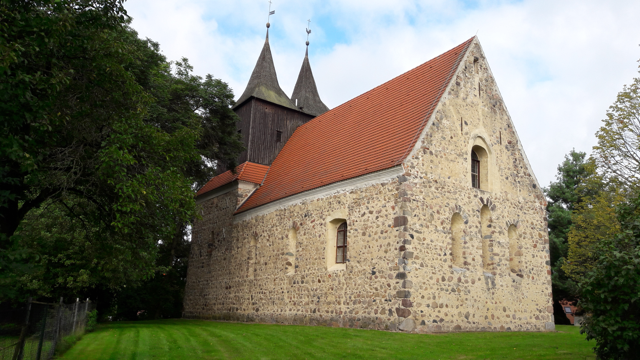 Auf einer Wiese steht die Dorchkirche Möbiskruge, umgeben von Bäumen, mit rotem Dach und hellem Steinmauerwerk im sommerlichen Licht.
