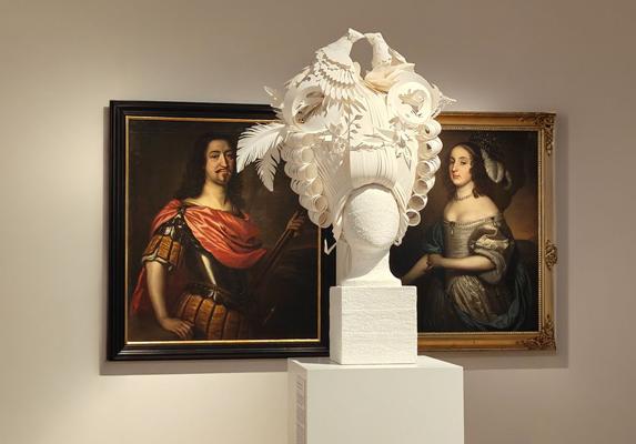 Eine filigrane weiße Papier-Skulptur steht auf einem Sockel vor zwei gerahmten historischen Porträts an einer Museumswand.