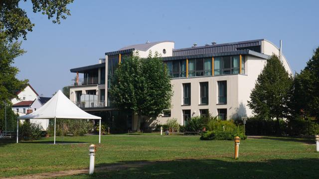 Vor dem modernen Haus am Spreebogen liegt eine große Rasenfläche; daneben steht ein weißes Zelt zwischen Bäumen.