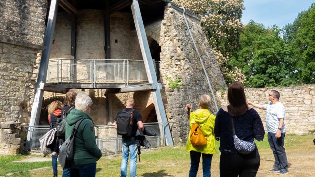Eine Besuchergruppe steht vor einer historischen Kalkofenanlage aus Stein und Metall und blickt zur Ruine.