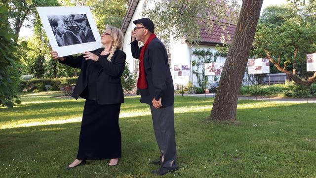 Eine blonde Frau und ein Mann in Mütze halten gemeinsam ein großes gerahmtes Schwarzweißfoto im Garten vor einem Haus.