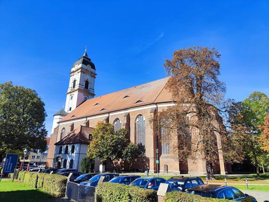 Eine große Backsteinkirche mit Turm und hohem Satteldach steht zwischen Bäumen; davor parken mehrere Autos im Sonnenschein.