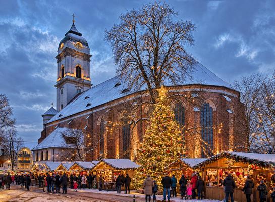 Vor der verschneiten Kirche stehen Weihnachtsmarktstände und ein leuchtender Baum, viele Menschen gehen vorbei.
