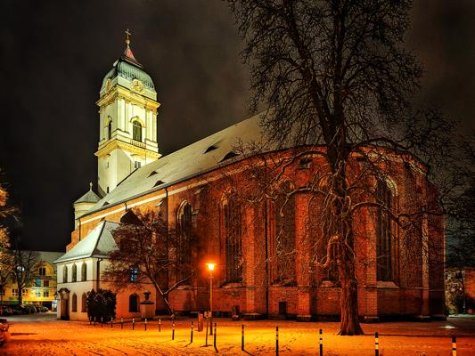 Dom St. Marien bei Nacht mit beleuchtetem Turm, roten Backsteinfassaden und verschneitem Vorplatz unter dunklem Himmel.