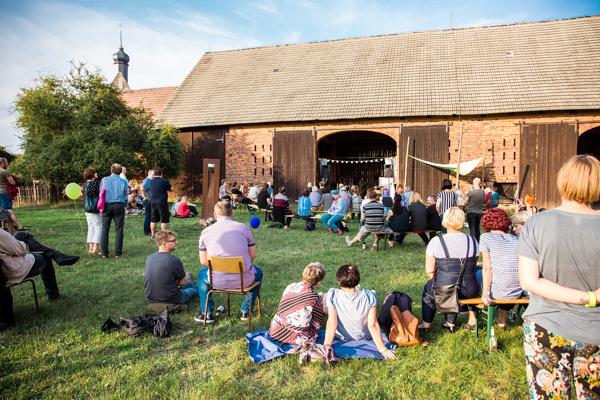 Bei der Eröffnung der openart Lausitz Biennale sitzen und stehen Besucher im Atelierhof Werenzhain vor der Scheune und verfolgen das Programm.