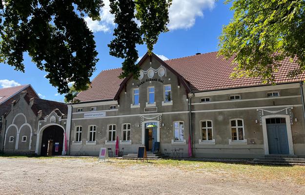 Vor dem Atelierhof Werenzhain zeigt sich das historische Gebäude der openart Lausitz Biennale Ausstellung mit rotem Dach und hellem Hof bei Sonnenschein.