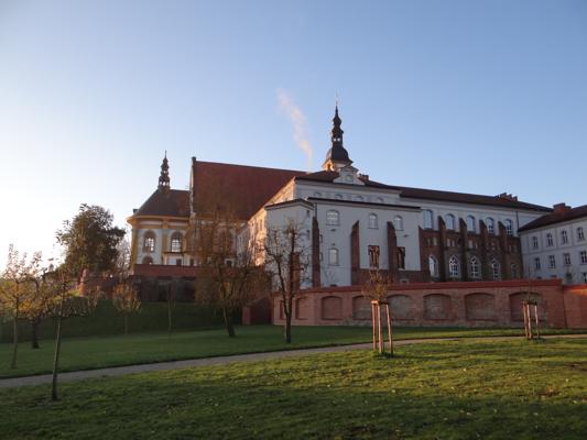 Die Klosteranlage zeigt lange barocke Fassaden, rote Dächer und einen Turm über einer grünen Wiese im Abendlicht.