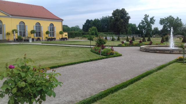 Ein weitläufiger Klostergarten mit gepflegten Beeten, Kieswegen und Springbrunnen liegt neben einem gelben Barockgebäude.