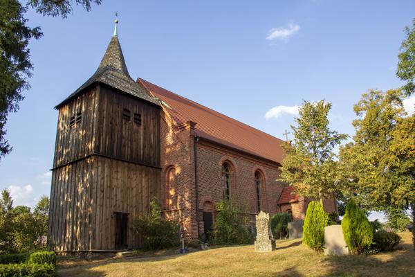 Zur SommerMusik in der Dorfkirche Grano zeigt sich die historische Kirche mit ihrem markanten Turm, rotem Dach und grüner Umgebung bei Sonnenschein.