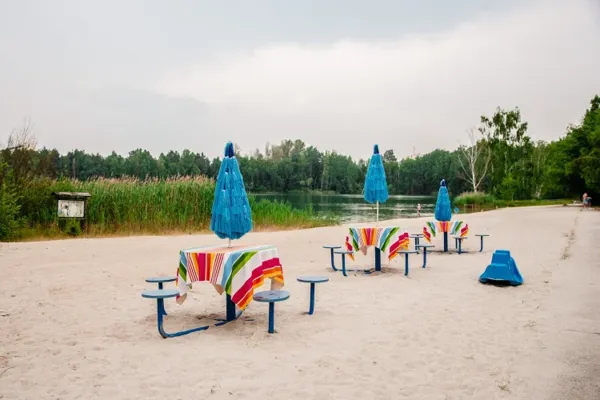 Am Rückersdorfer See stehen bunt gedeckte Tische unter blauen Schirmen für die openart Lausitz Biennale „Die Welt zu Gast“ auf dem Sandstrand.